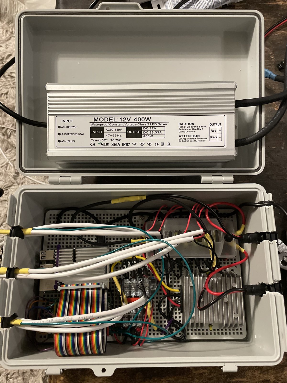 The open controller case on a workbench: a 12V 400W waterproof LED driver mounted in the lid, and in the body a Raspberry Pi with a rainbow ribbon cable, terminal blocks, a converter and the runs leaving through sealed glands in the wall