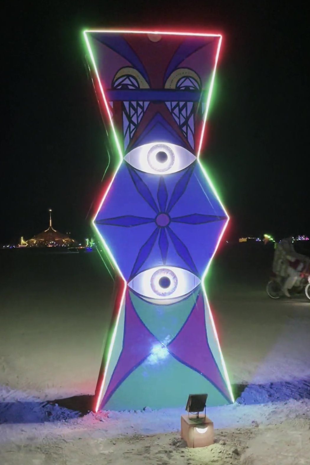 The pillar lit at night on the open playa, its edges outlined in green and red light, two large eyes glowing on the painted face, a spotlight at its foot and the lights of Black Rock City low on the horizon