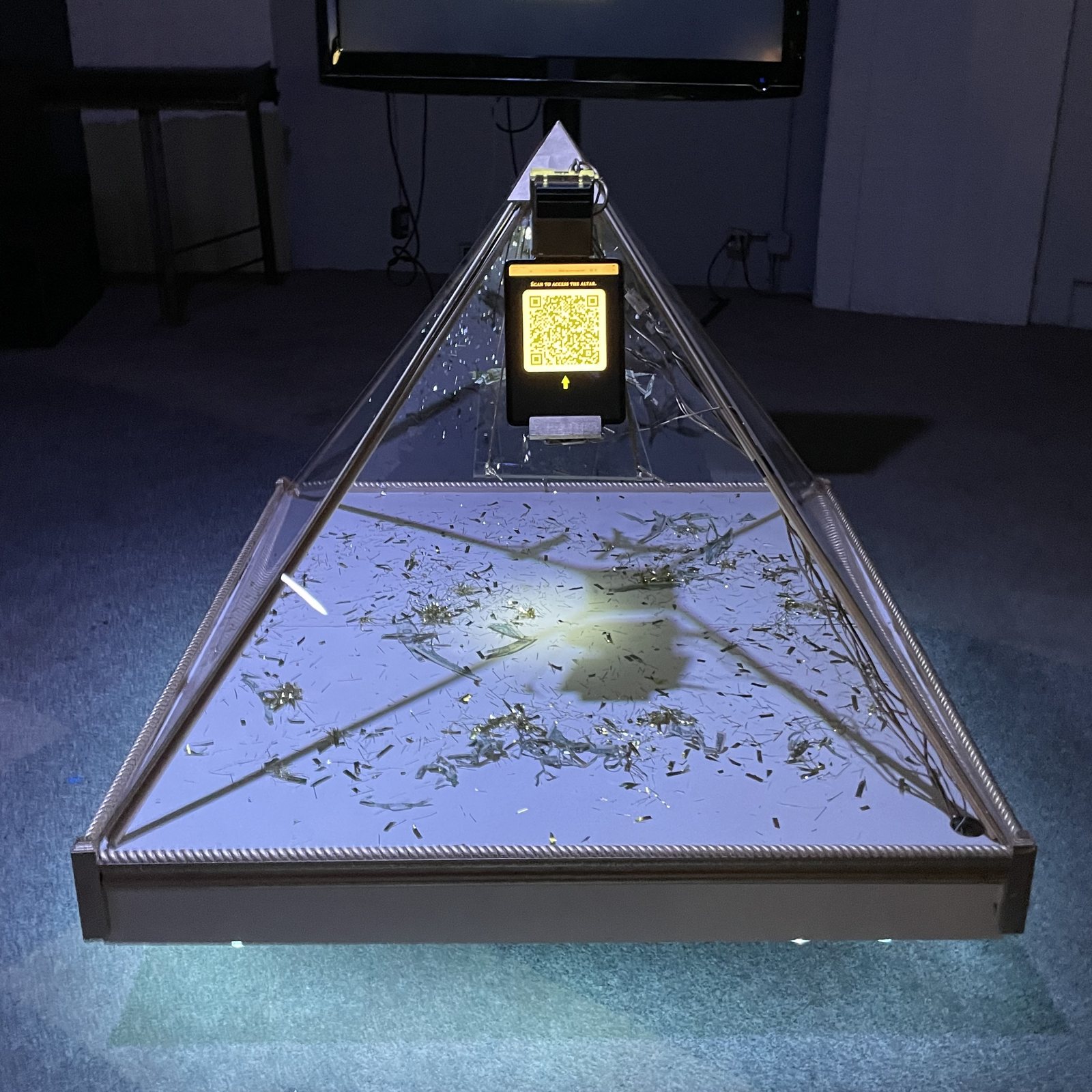 A lit glass pyramid on a dark gallery floor, a glowing QR screen at its apex and shredded banknotes on the platform inside it