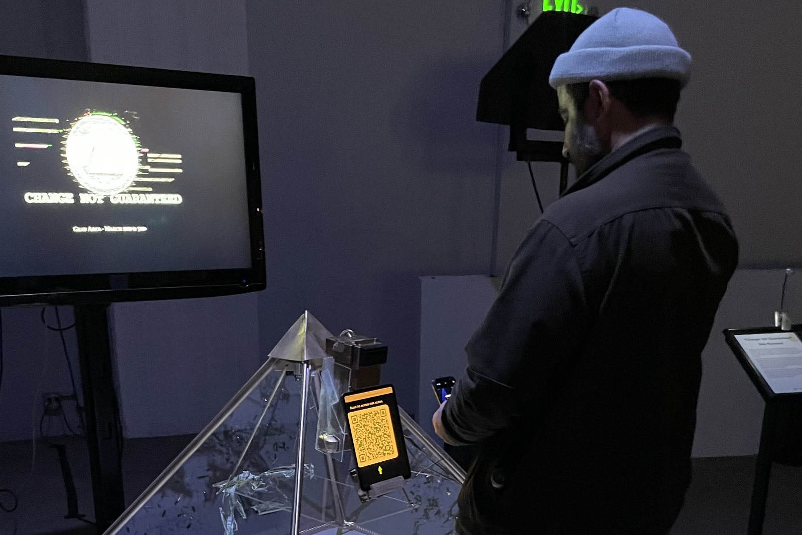 A visitor in a wool cap standing over the pyramid with a phone in hand, the gallery screen beside them showing the token and the title Change Not Guaranteed