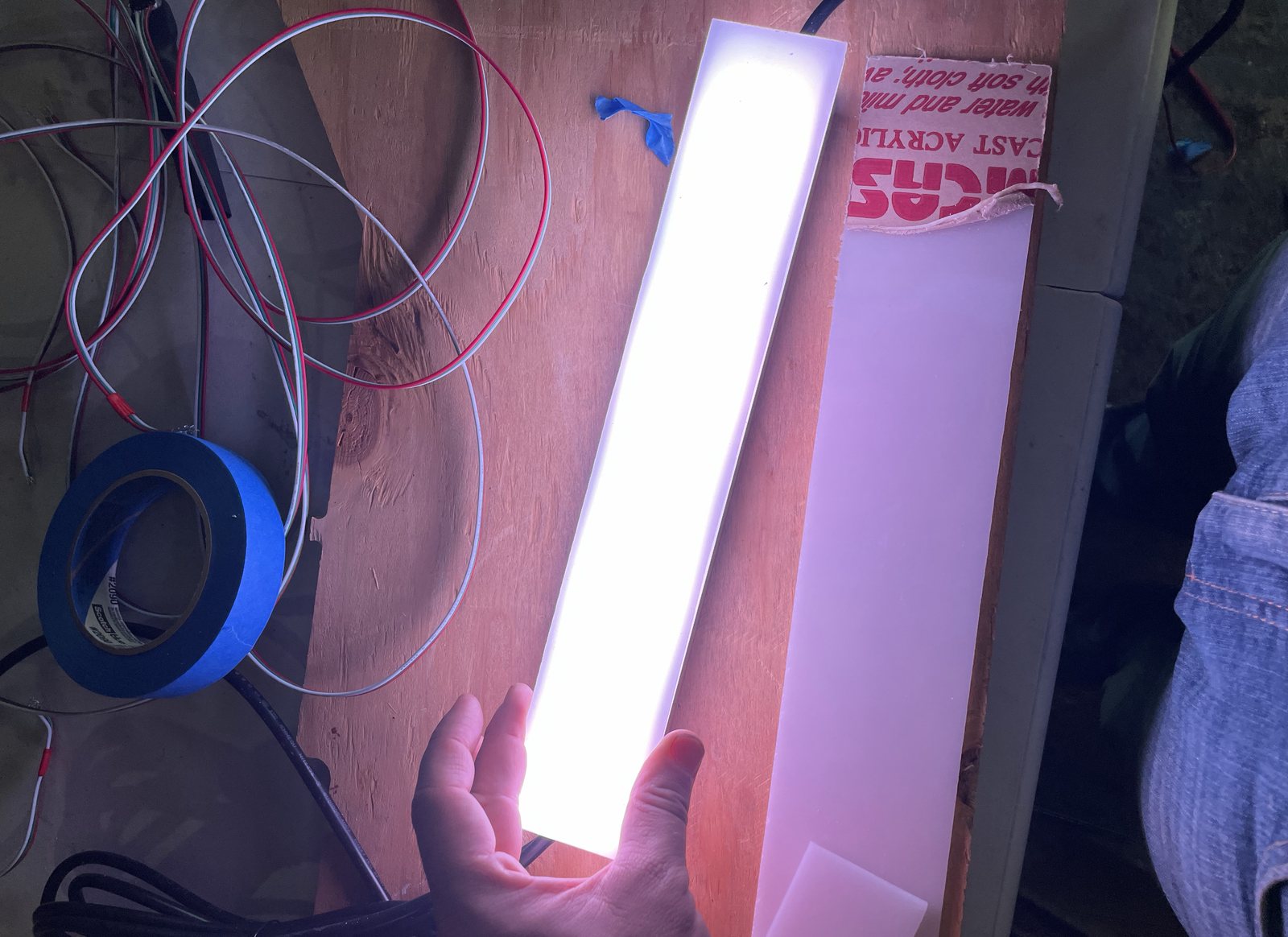 A custom fin light held over a workbench, a long strip of cast acrylic glowing white and pink, with wire and a roll of tape beside it and an uncut sheet of the same acrylic alongside