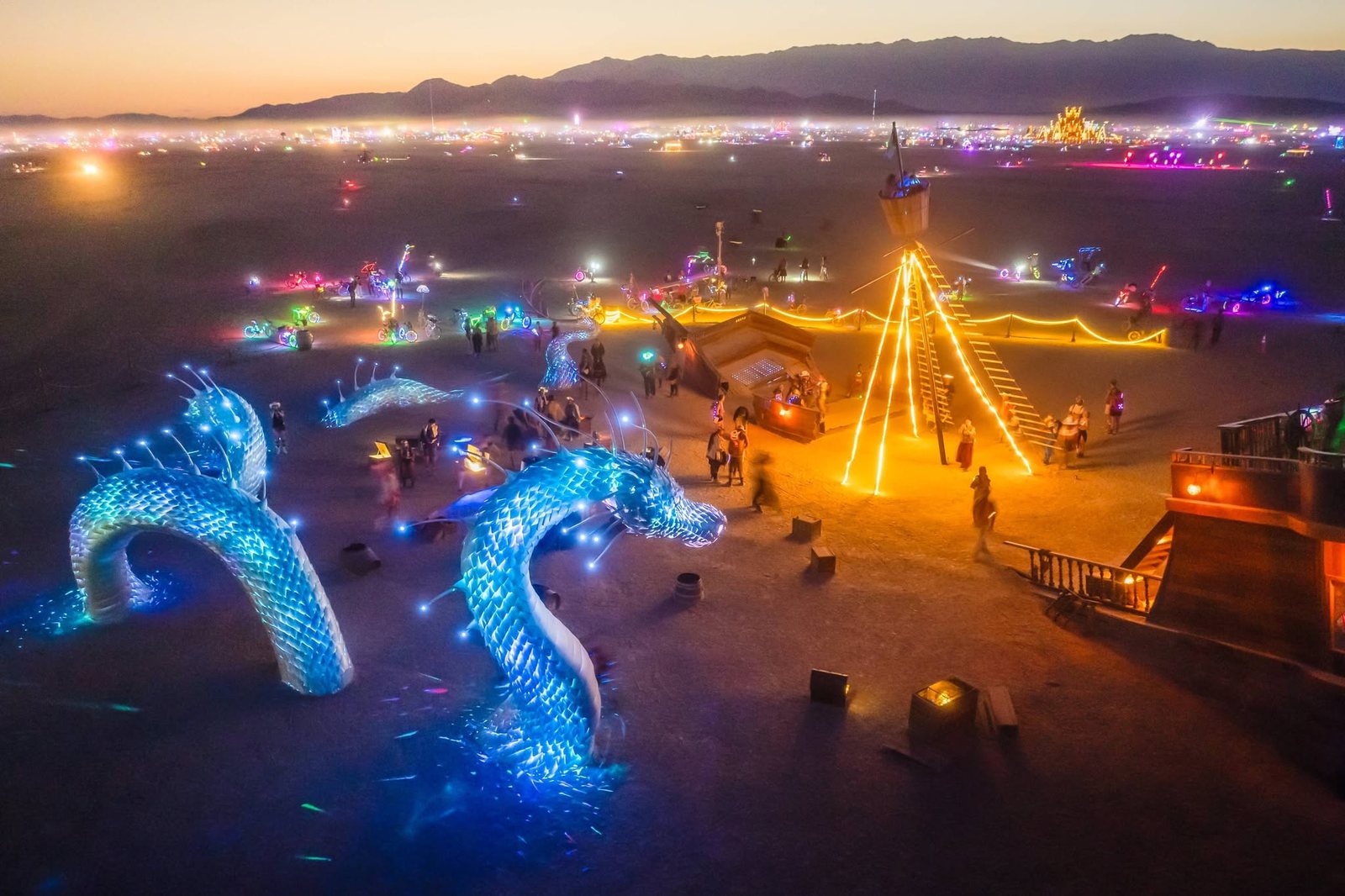 The installation at dusk from above: Naga lit blue rising out of the open playa, the Captainess and her lit mast to the right, and Black Rock City glowing along the horizon