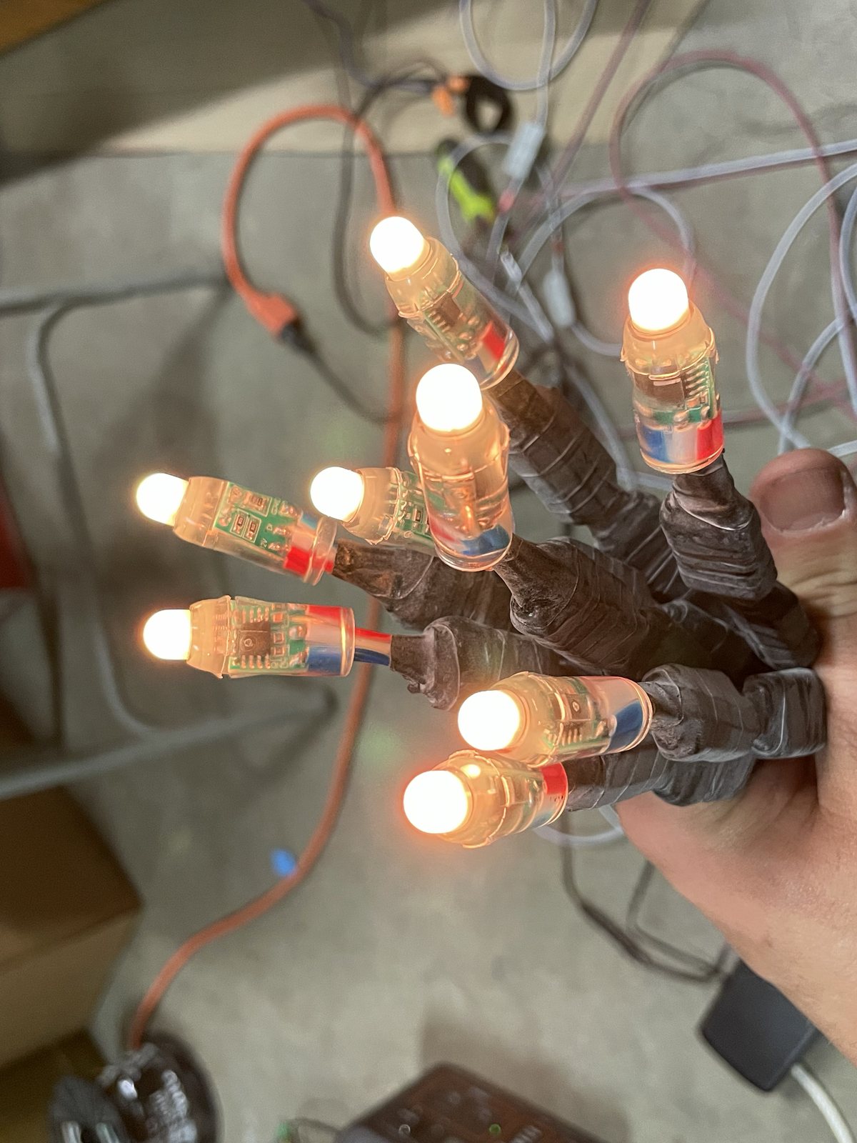 A hand holding a bundle of bare addressable pixel nodes, each lit warm orange, their circuit boards visible through clear plastic caps and their leads taped into a trunk