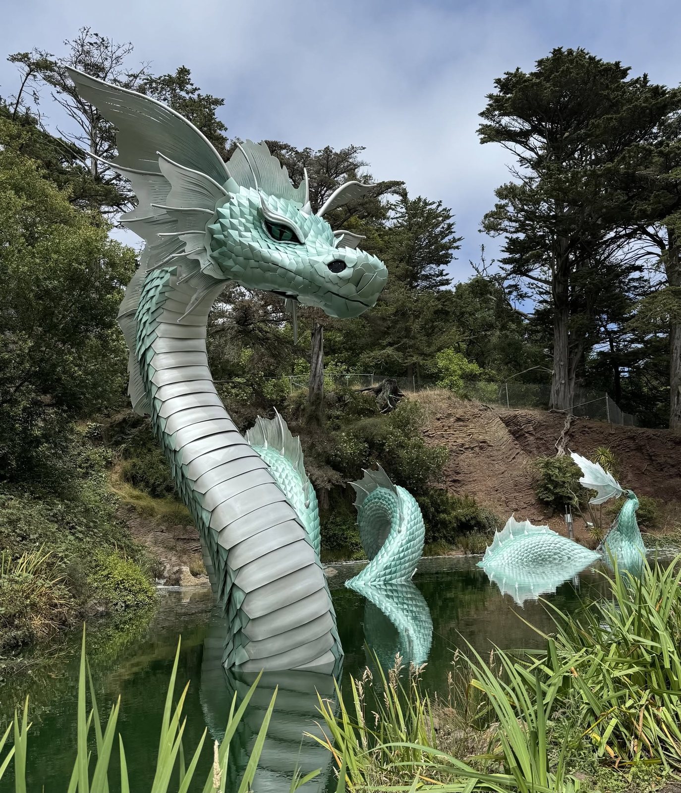 Naga in daylight from the path, pale green scales and fins rising out of the pond with trees behind