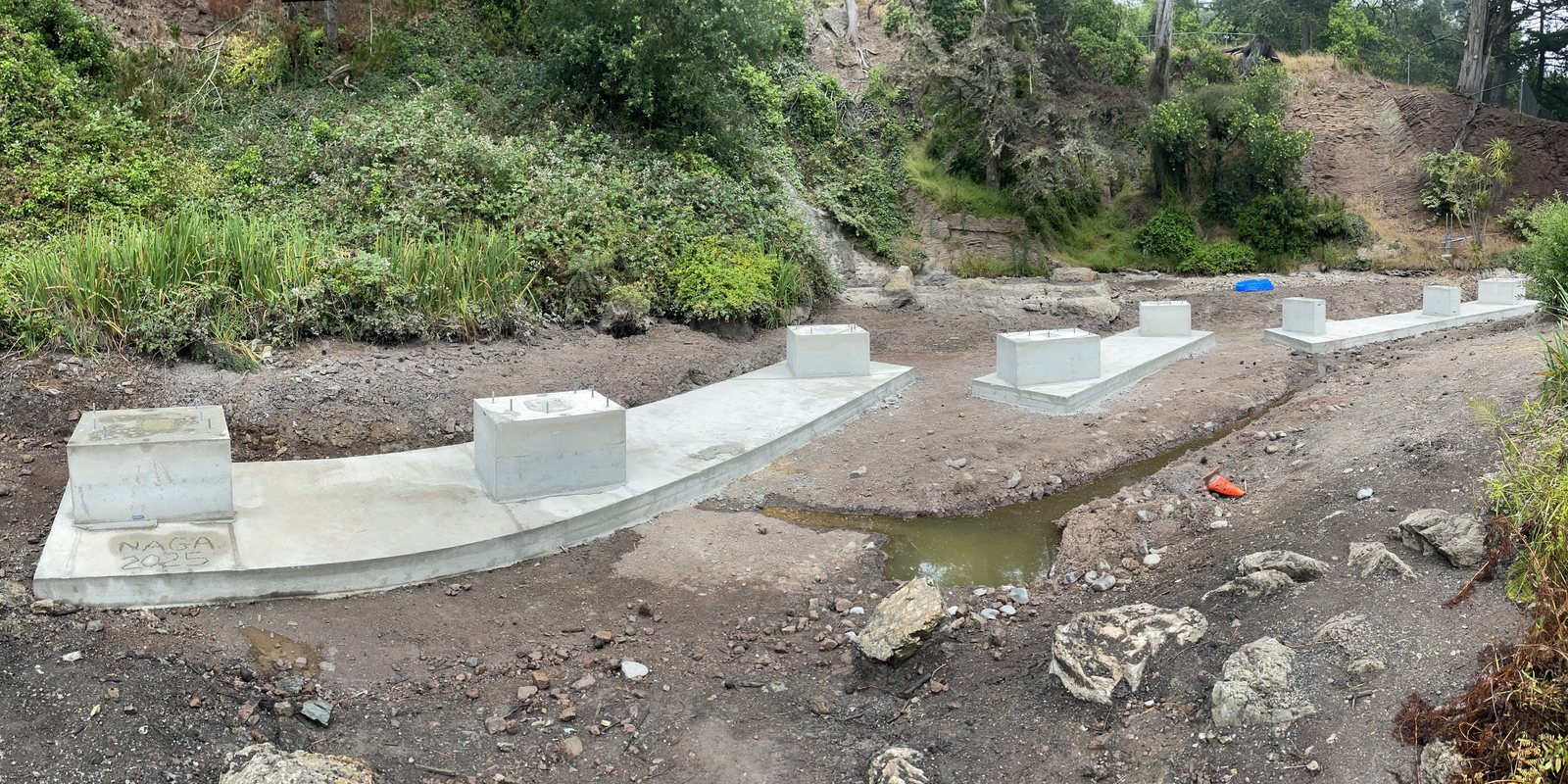 The drained pond with a curving line of fresh concrete footings poured along its bed, NAGA 2025 scratched into the nearest one