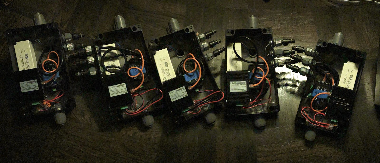 Five black NEMA enclosures laid out in a row on a wood floor, each wired with a Meanwell power supply, a controller and sealed cable glands