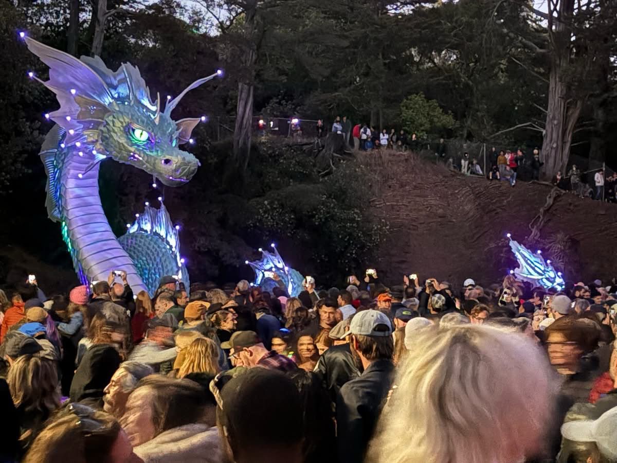 A dense opening night crowd at dusk holding up phones toward Naga, his head and fins lit violet above them